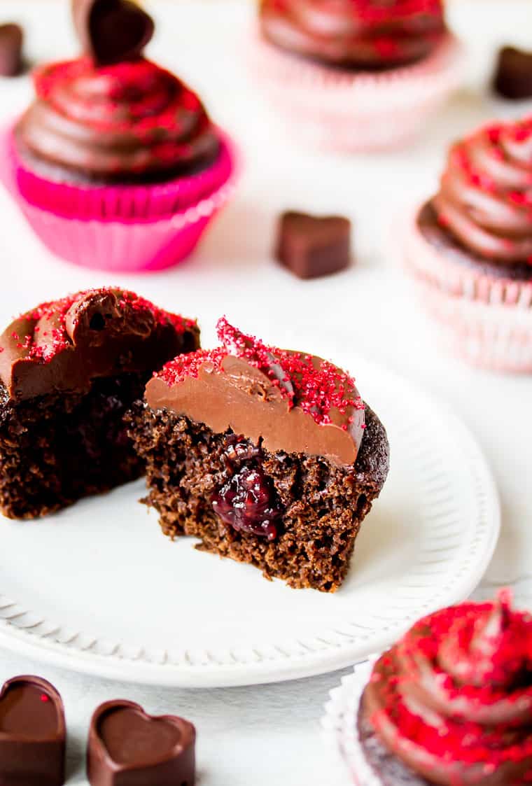 A chocolate cupcake cut in half to show raspberry jam filling on a white plate with more cupcakes and heart shape chocolate candy around it