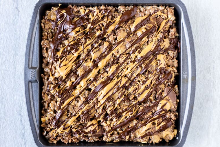 Chocolate Peanut Butter Rice Krispies Treats with Reese's cups and melted chocolate and peanut butter drizzle on top in a square baking dish over a white background