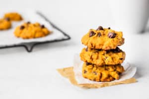 3 Pumpkin chocolate chip cookies stacked on top of each other with a mug and cooling rack with more cookies on it in the background