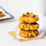 3 pumpkin chocolate chip cookies stacked on top of each other with part of a cooling rack and a mug in the background
