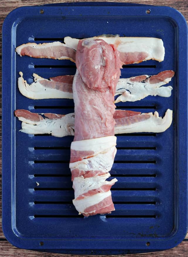 Pork tenderloin being wrapped with bacon on a blue broiler pan