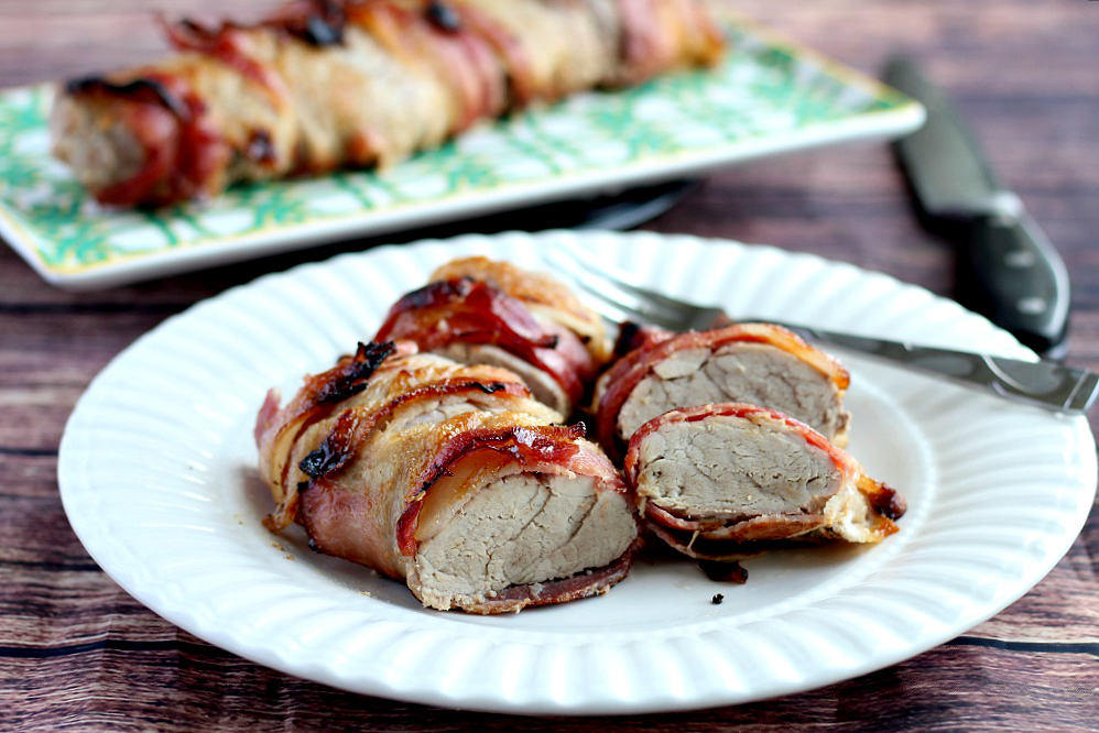 Bacon Wrapped Pork sliced on a white plate with a serving tray of more pork in the background on a wood backdrop