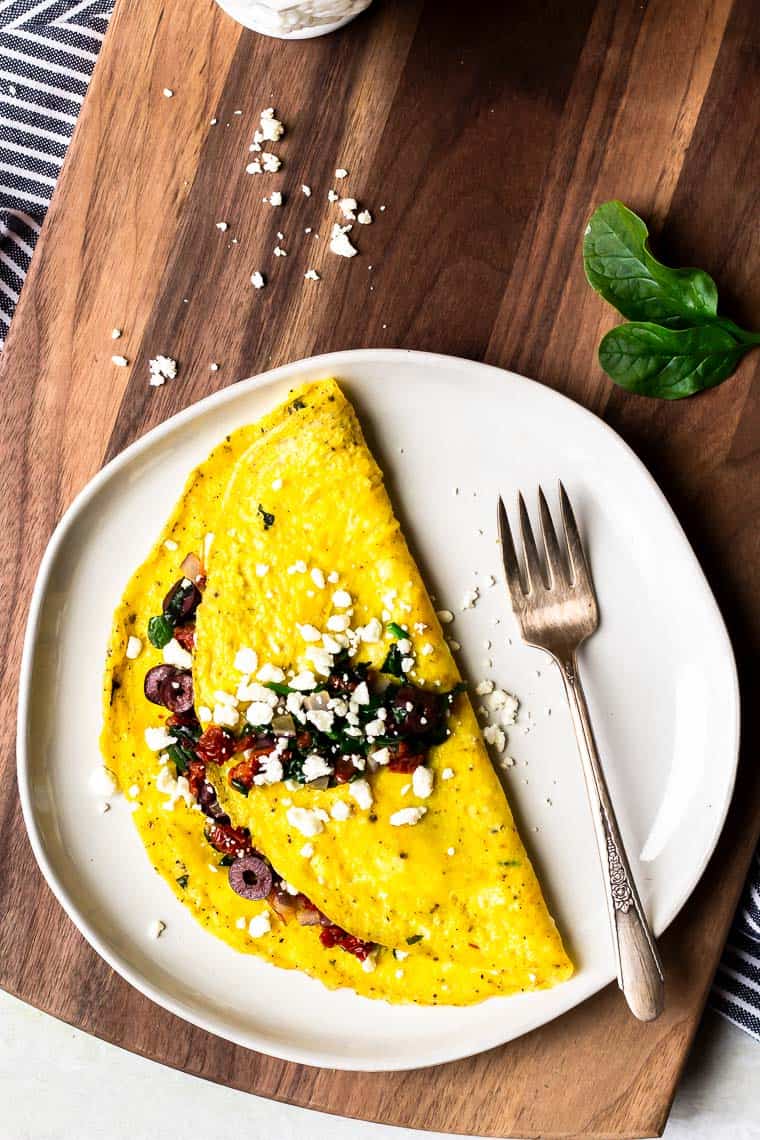 Overhead of a Greek Omelet on a white plate with a fork over a wood board with feta cheese, spinach, a white cup, and a blue and white towel in the background