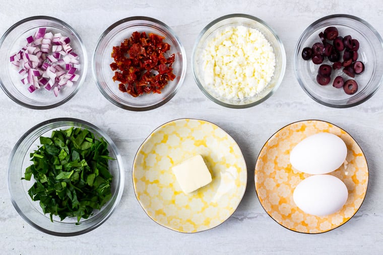 Ingredients needs to make a Greek Omelet in glass bowls and small plates over a white background