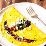A Greek Omelet on a white plate with a fork on a wood board with feta cheese and spinach in the background