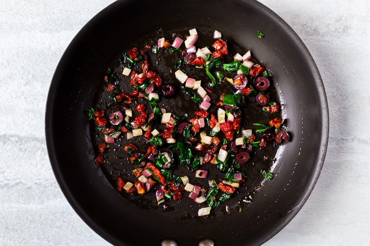 Spinach, sun-dried tomatoes, red onion, and olives cooking in a black skillet over a white background