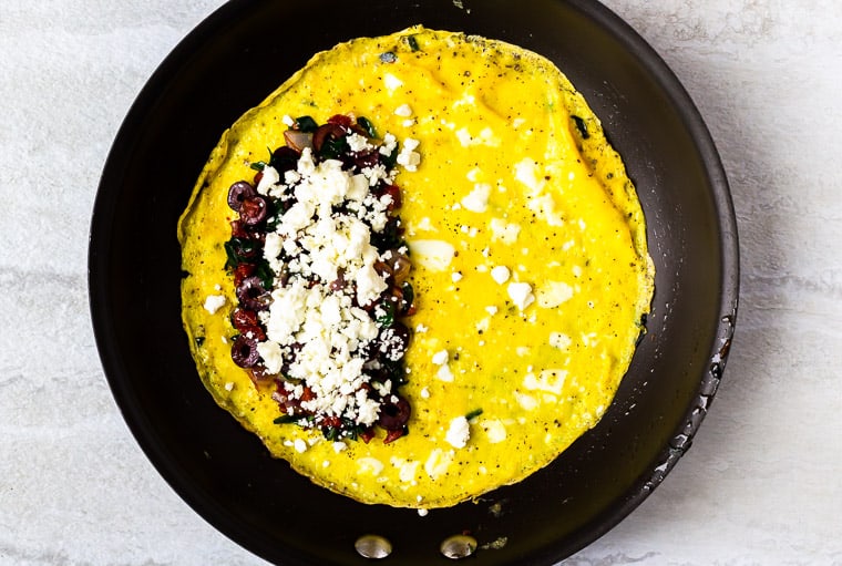An omelet in a black skillet topped with spinach, sun-dried tomatoes, red onion, olives, and feta cheese over a white background
