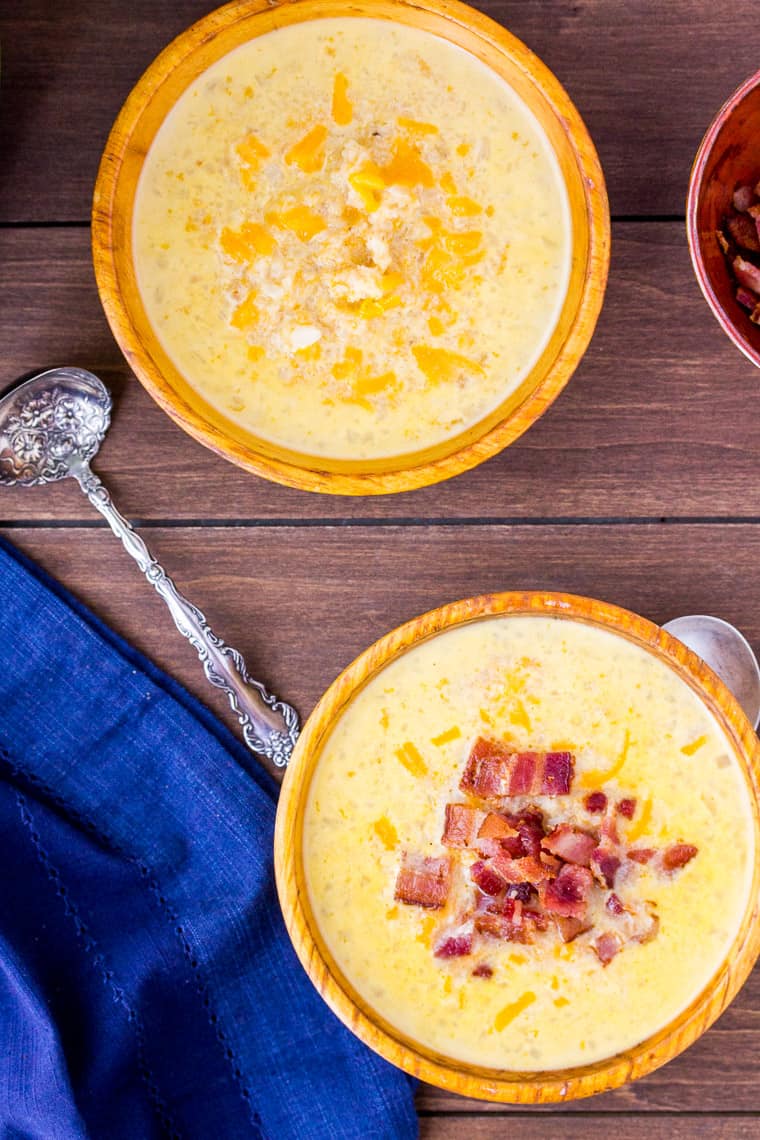 Cheesy Cauliflower Soup in 2 wood bowls with 2 spoons. One bowl of soup has bacon on top. over a wood backdrop