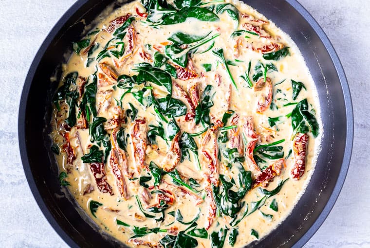 A skillet with spinach and sun-dried tomatoes in a cream sauce over a white background