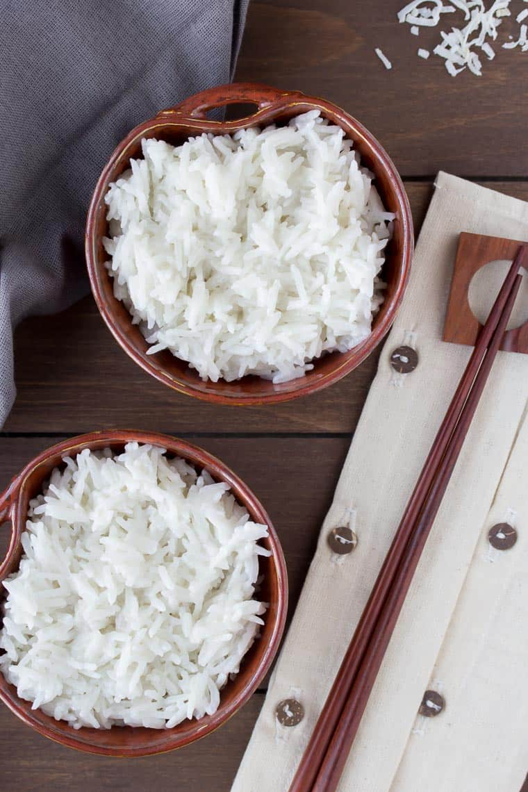 Overhead of 2 brown bowls filled with coconut rice on a wood background with a gray napkin and chopsticks