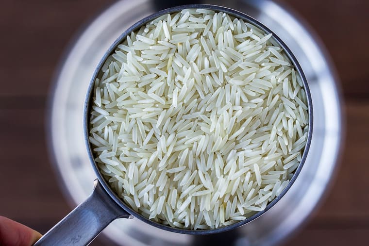 Close up of rice in a measuring cup being held up over a silver pot on a wood background