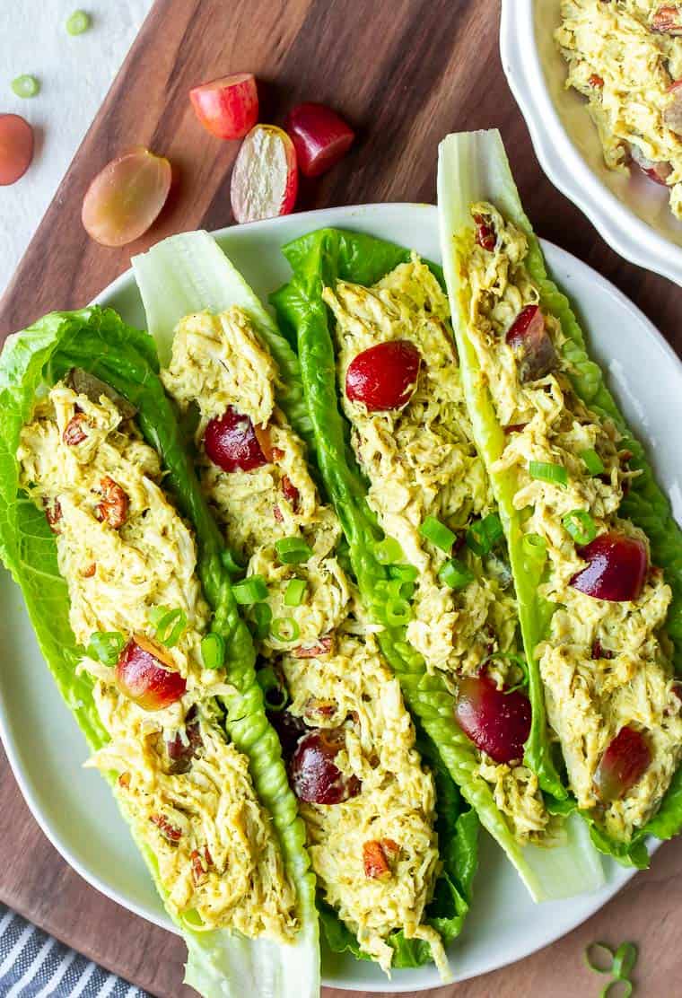 4 lettuce leaves filled with Curry Chicken Salad on a white plate over a wood board with grapes and a part of a bow with more chicken in it in the background
