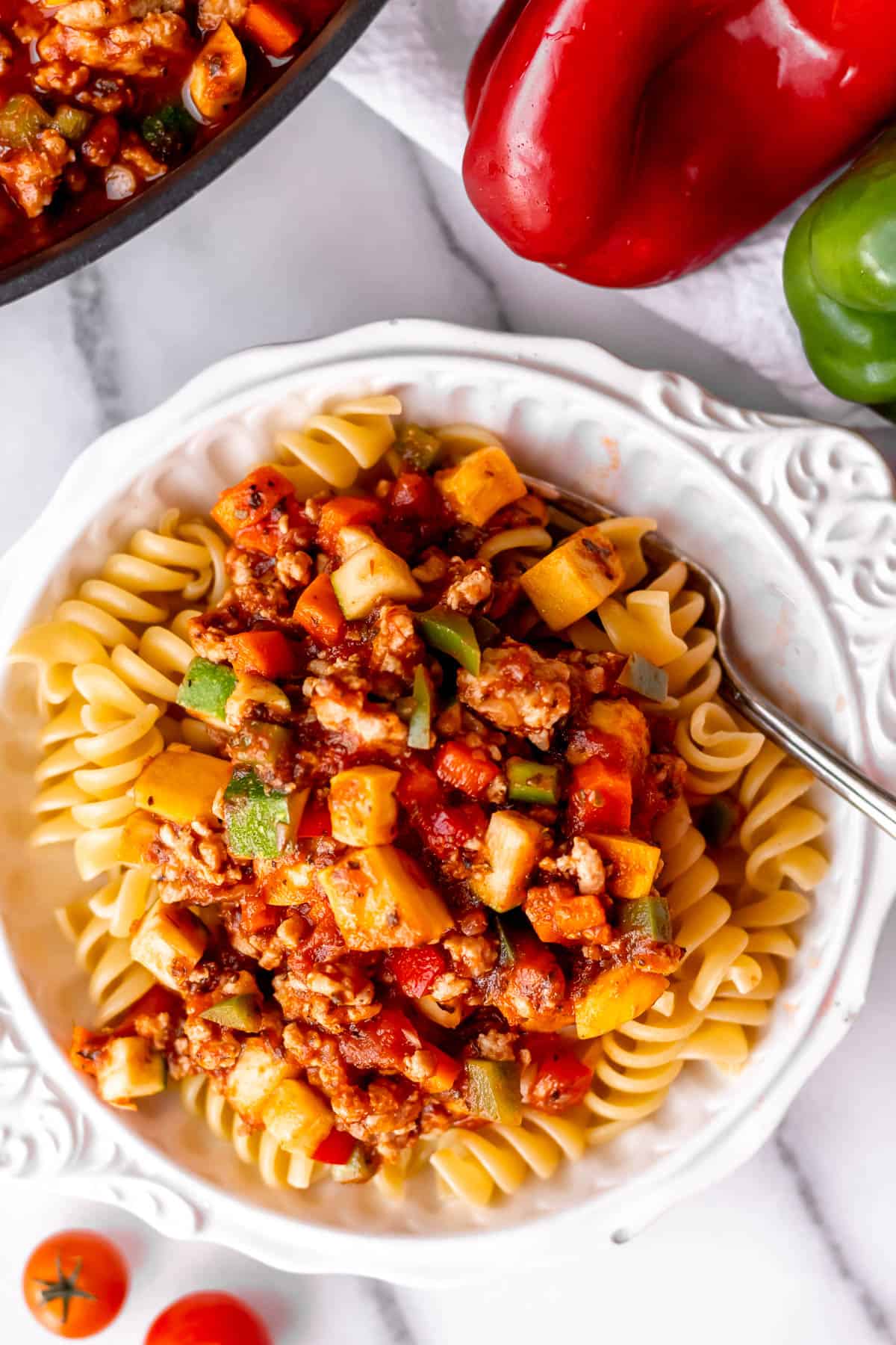 Pasta and vegetable spaghetti sauce in a white bowl with peppers and part of the skillet with more sauce in it partially showing.