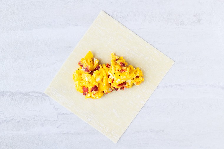 An egg rolls wrapper with bacon and cheese scrambled eggs in the center on a white background
