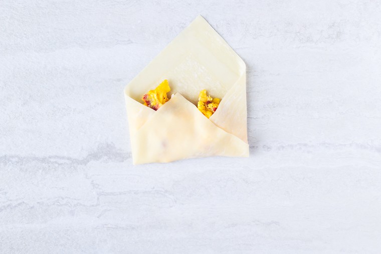 An egg roll wrapper with 3 corners folded in over scrambled eggs on a white background