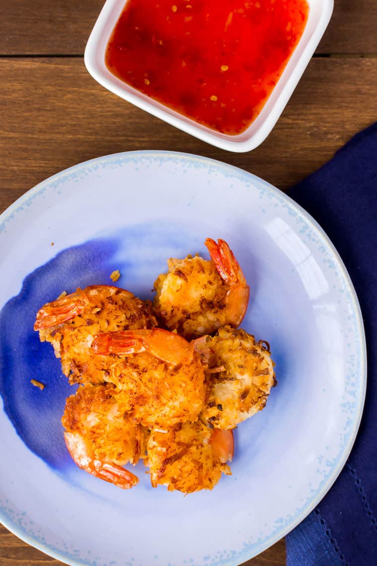A blue plate of gluten free coconut shrimp with a white, square bowl of sweet chili sauce on wood backdrop with a blue napkin