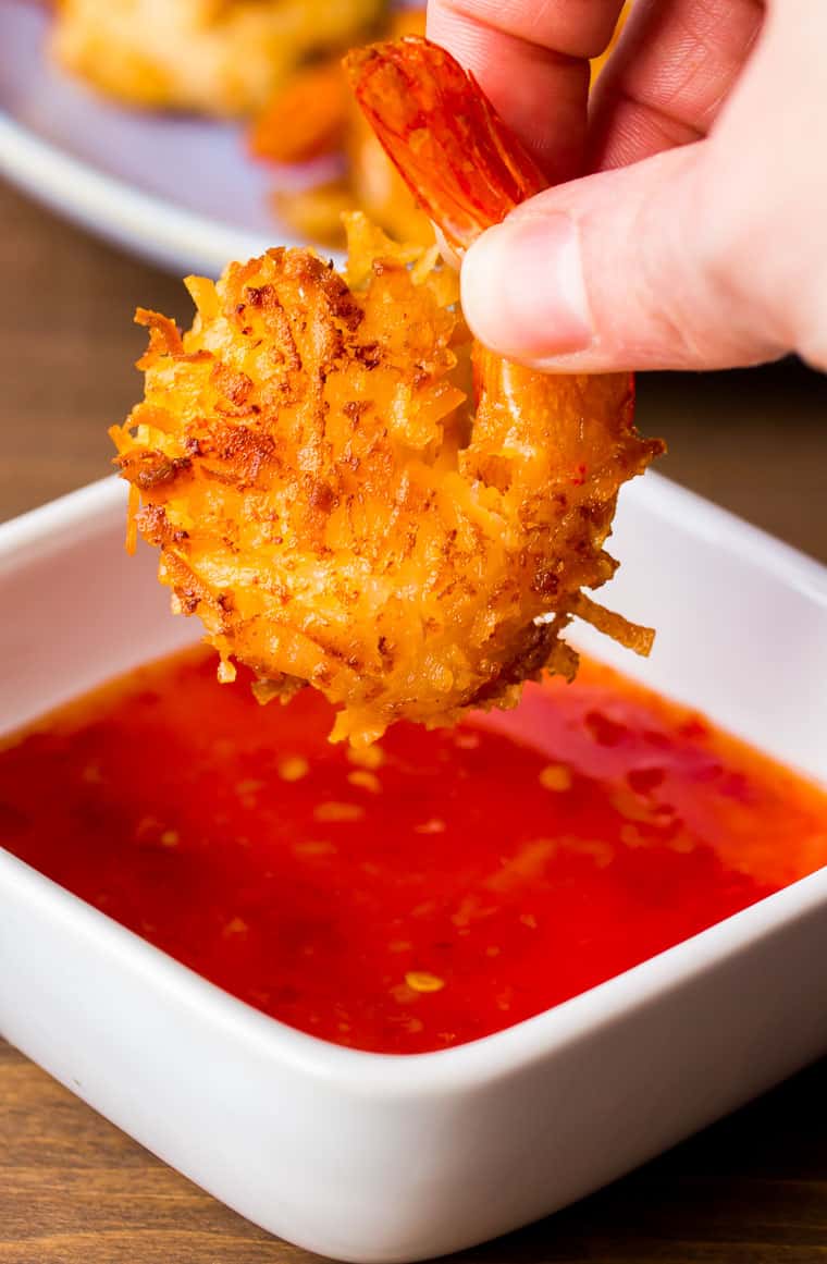 Close up of coconut shrimp about to be dipped in sweet chili sauce in a white bowl on a wood table