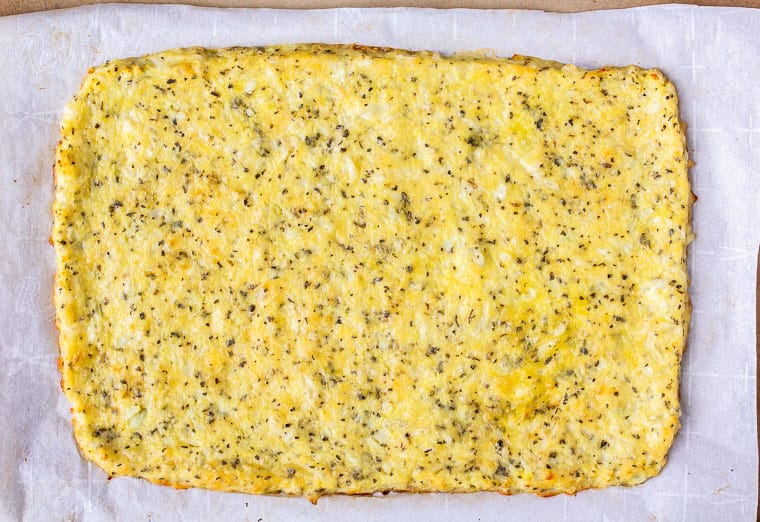 A rectangular baked cauliflower crust on parchment paper on a pizza stone