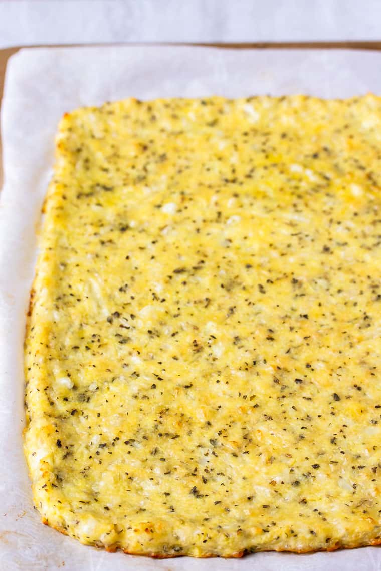 Close up of a rectangular baked cauliflower pizza crust on parchment paper on a pizza stone