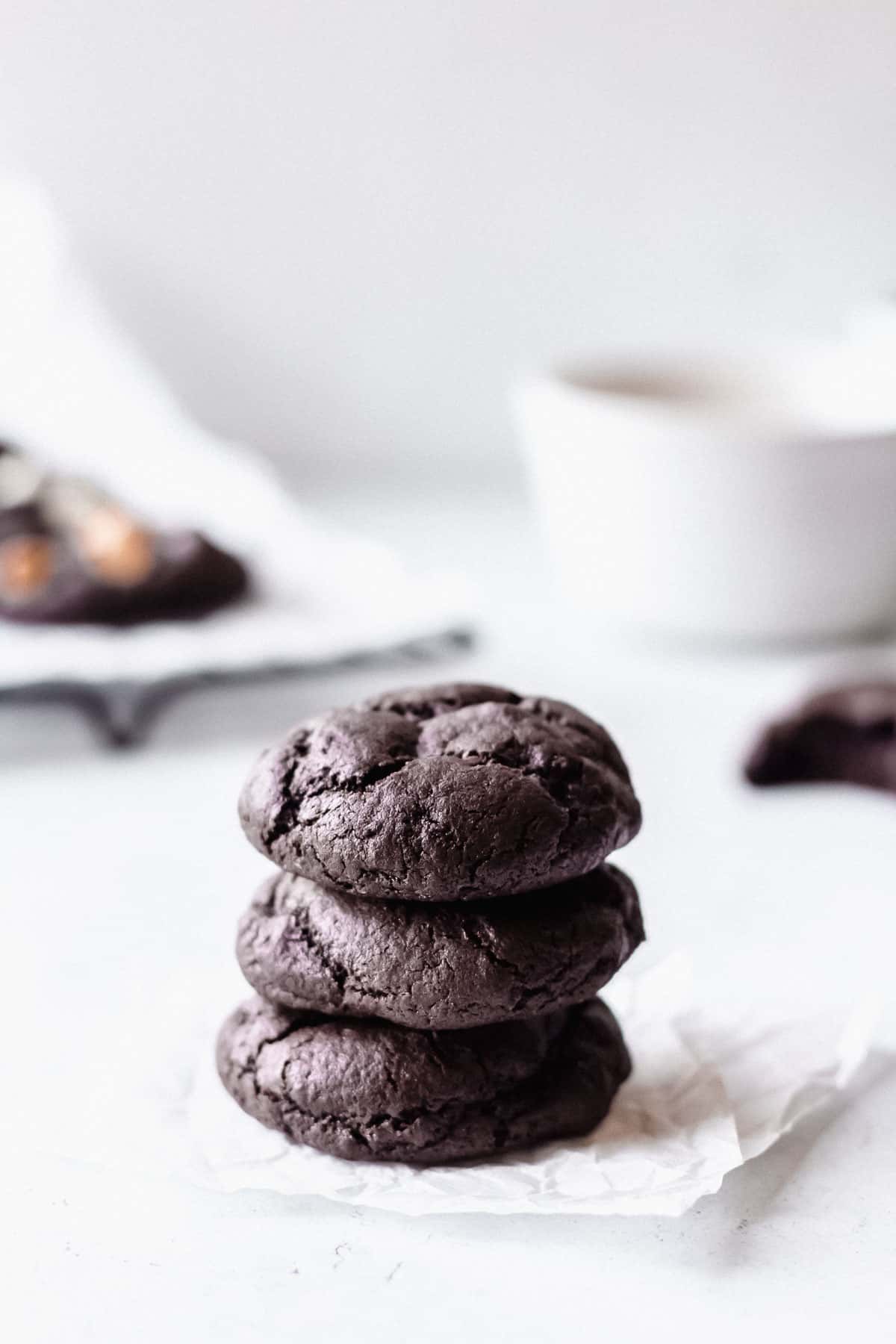 3 dark chocolate cookies stacked on top of each other with more cookies and a white bowl blurred in the background