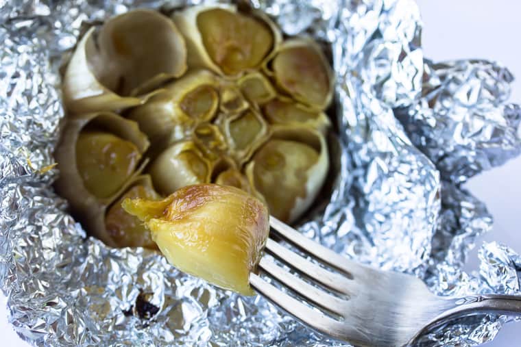 A fork with a roasted garlic clove in front of a bulb of roasted garlic in foil
