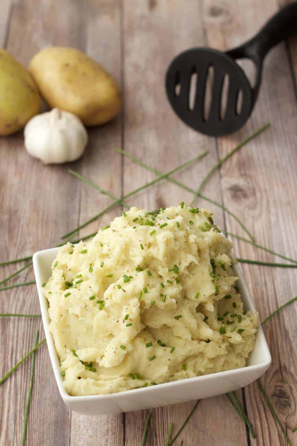 Vegan Garlic Mashed Potatoes in a white square dish with potatoes, garlic, and a black spoon in the background