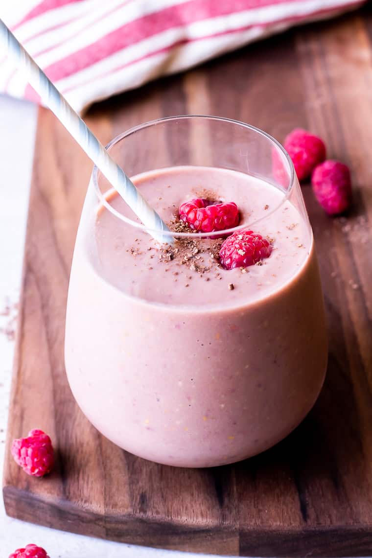 Chocolate raspberry smoothie in a glass with a straw on a wood board with extra raspberries and a white and red striped towel in the background