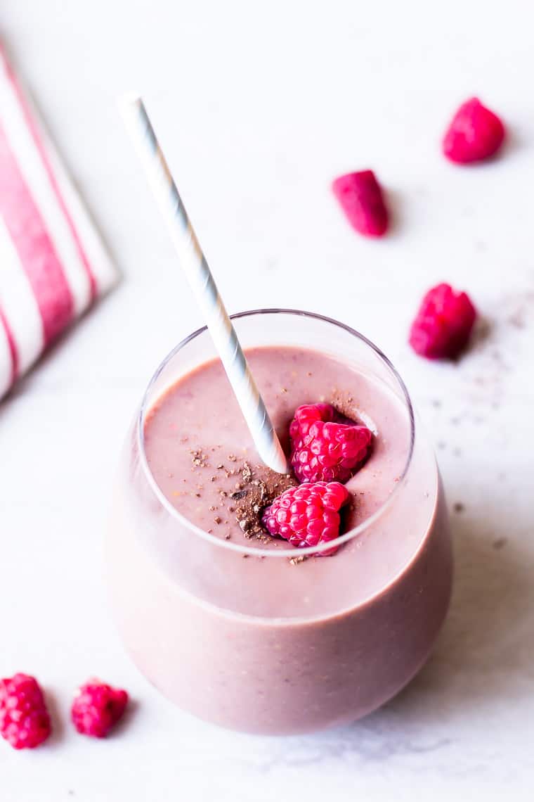 A chocolate raspberry smoothie in a glass with a straw on a white background with 5 extra raspberries and a red striped towel blurred in the background