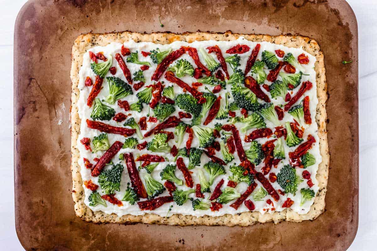 A pizza crust topped with ricotta cheese, broccoli and sun-dried tomatoes