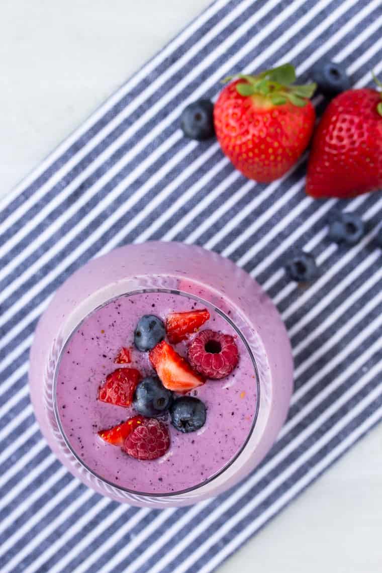 Overhead of a Mixed berry smoothie topped with fresh berries over a blue and white striped napkin with more strawberries and blueberries in the background