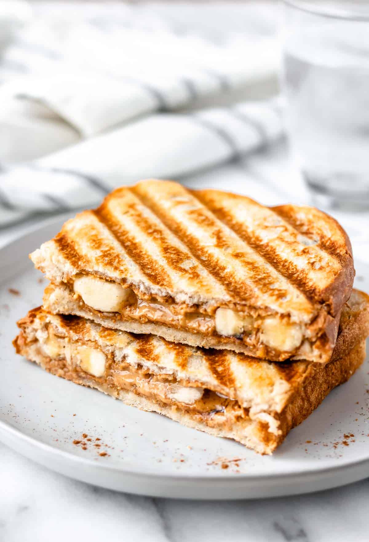 Two halves of a fried peanut butter and banana sandwich on a white plate with a glass of water and towel in the background.