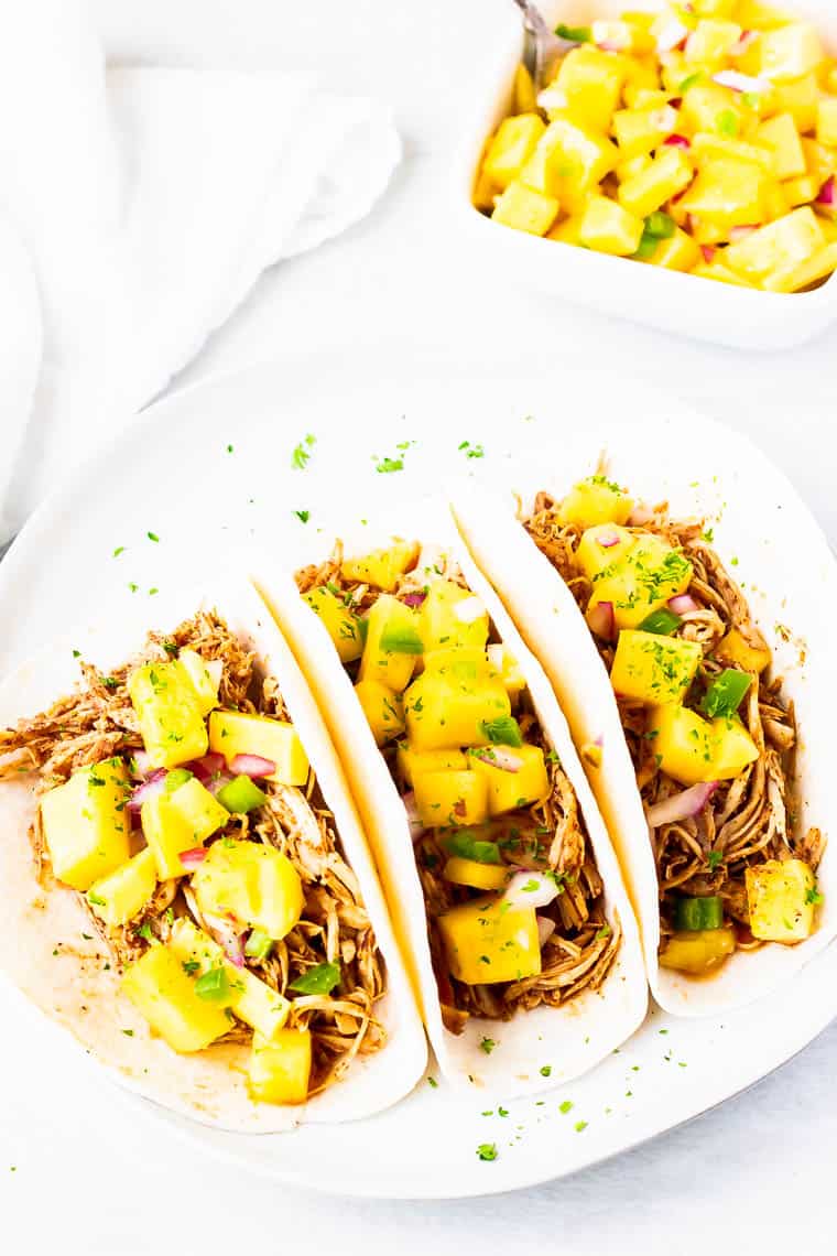 3 Shredded Chicken Tacos topped with diced mango salsa on a white plate over a white background with a white napkin and white bowl of mango salsa in the background
