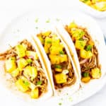 3 Shredded Chicken Tacos topped with diced mango salsa on a white plate over a white background with a white bowl of mango salsa in the background