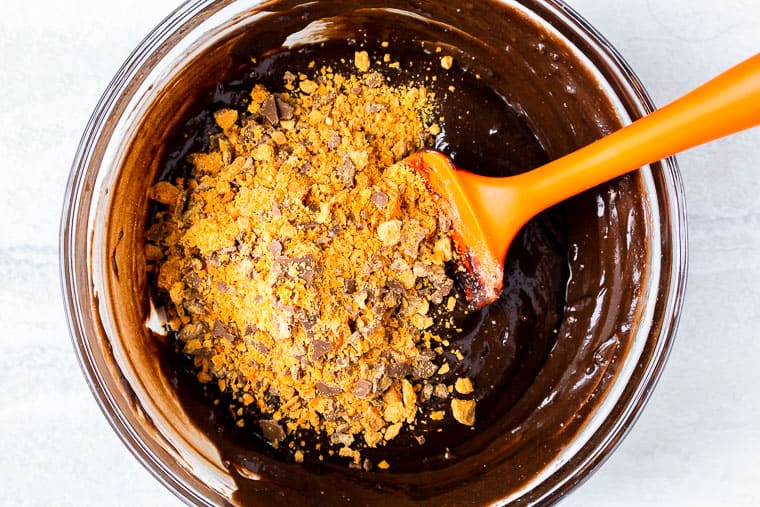 Brownie mix with crushed Butterfinger bars on top and an orange spatula over a white backdrop