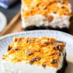 Close up of a Butterfinger Cheesecake Bar on a white plate with a second one in the background with a fork and napkin