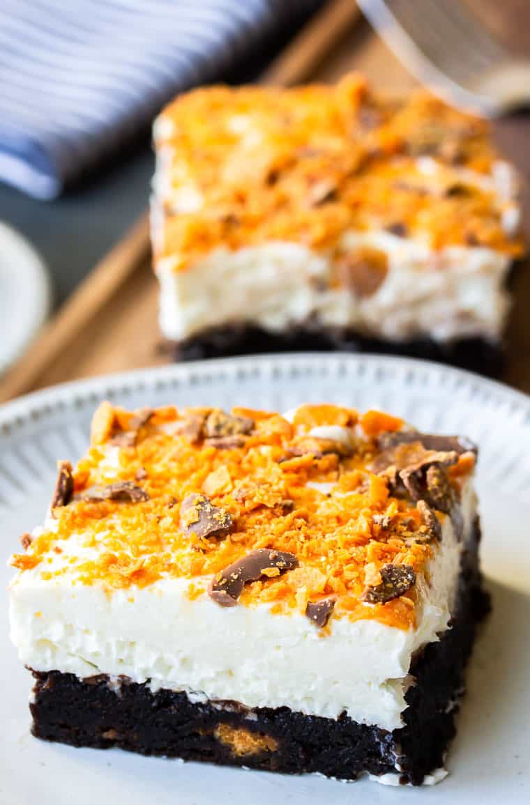 Close up of a Butterfinger Cheesecake Bar on a white plate with a second one in the background with a fork and napkin