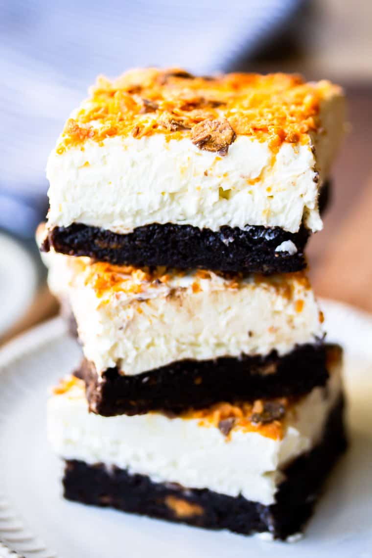 2 Butterfinger Cheesecake Bars stacked on top of each other on a white plate over a wood board with a blurred cloth napkin and fork in the background