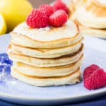 A Stack of Lemon Ricotta Pancakes on a Blue Plate with Raspberries and Lemons in the Background