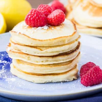 A Stack of Lemon Ricotta Pancakes on a Blue Plate with Raspberries and Lemons in the Background