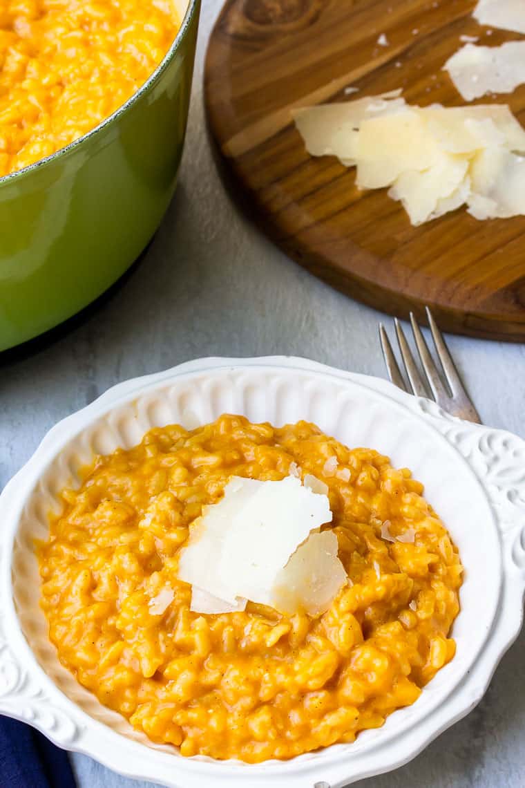 Pumpkin risotto in a white bowl with a green dutch oven with more risotto it and a wood board with cheese shavings in the background