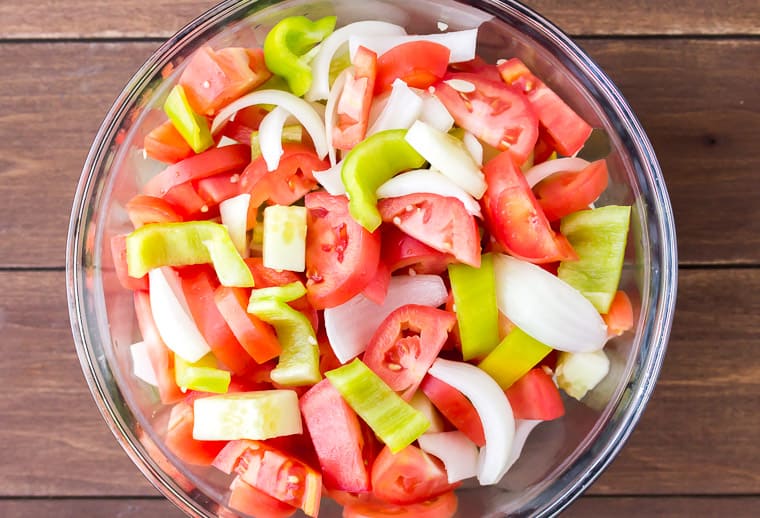 Tomatoes, peppers, onion, and cucumber chunks in a glass bowl over a wood background