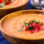 Close up of gazpacho soup in an orange wood bowl on a dark blue napkin with a second bowl and 2 spoons in the background