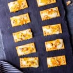 Apricot Jam Crackers with Asiago and Walnuts on a slate board over a blue background with a blue and white towel and a spoon of apricot preserves in the background
