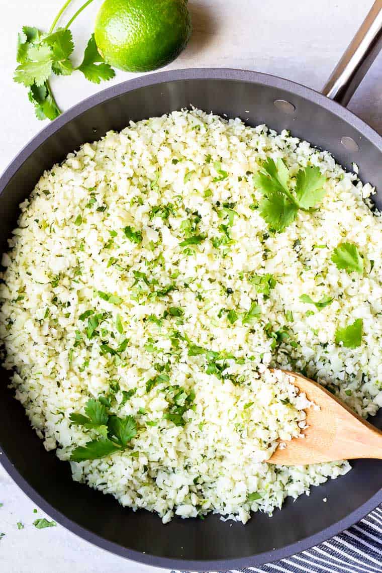 overhead of cilantro lime cauliflower rice with cilantro and lime with a wood spoon in it and cilantro and a lime in the background