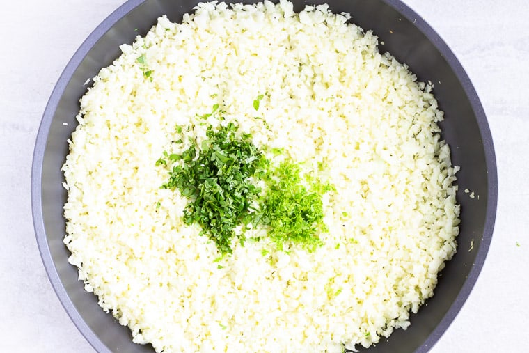 Cilantro and lime zest on top of the cauliflower rice in a black skillet over a white background
