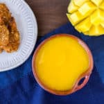 Sweet Mango Dipping Sauce in a dark orange bowl with a blue napkin, a white plate of shrimp, fresh mango, and limes in the background