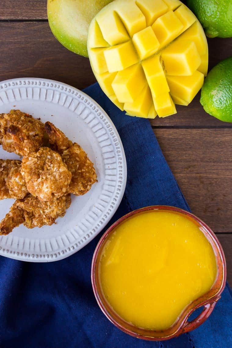 Overhead view of sweet mango dipping sauce with shrimp on a white plate and half a mango and 2 limes in the background all on a wood backdrop