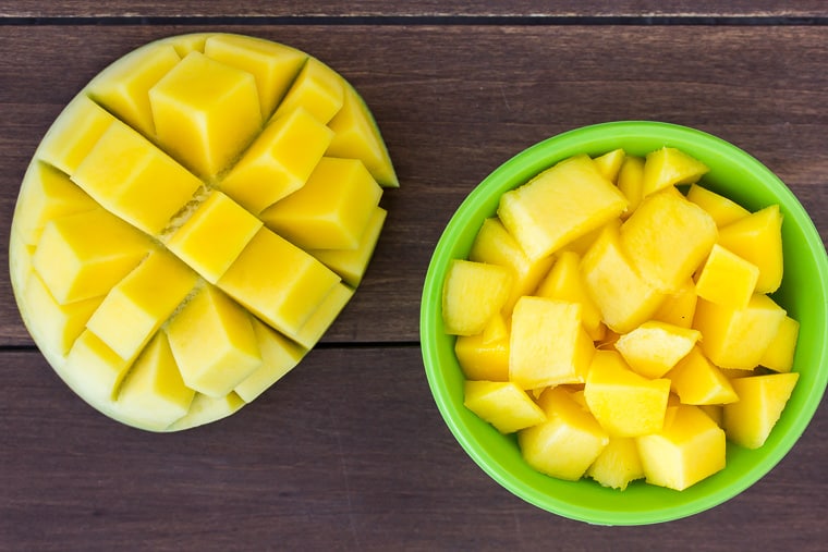Diced mango in a green bowl with a half of a mango on the side on a wood backfground