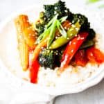 Vegetable stir fry on rice in a white bowl over a white background with a white napkin and slice of green onion