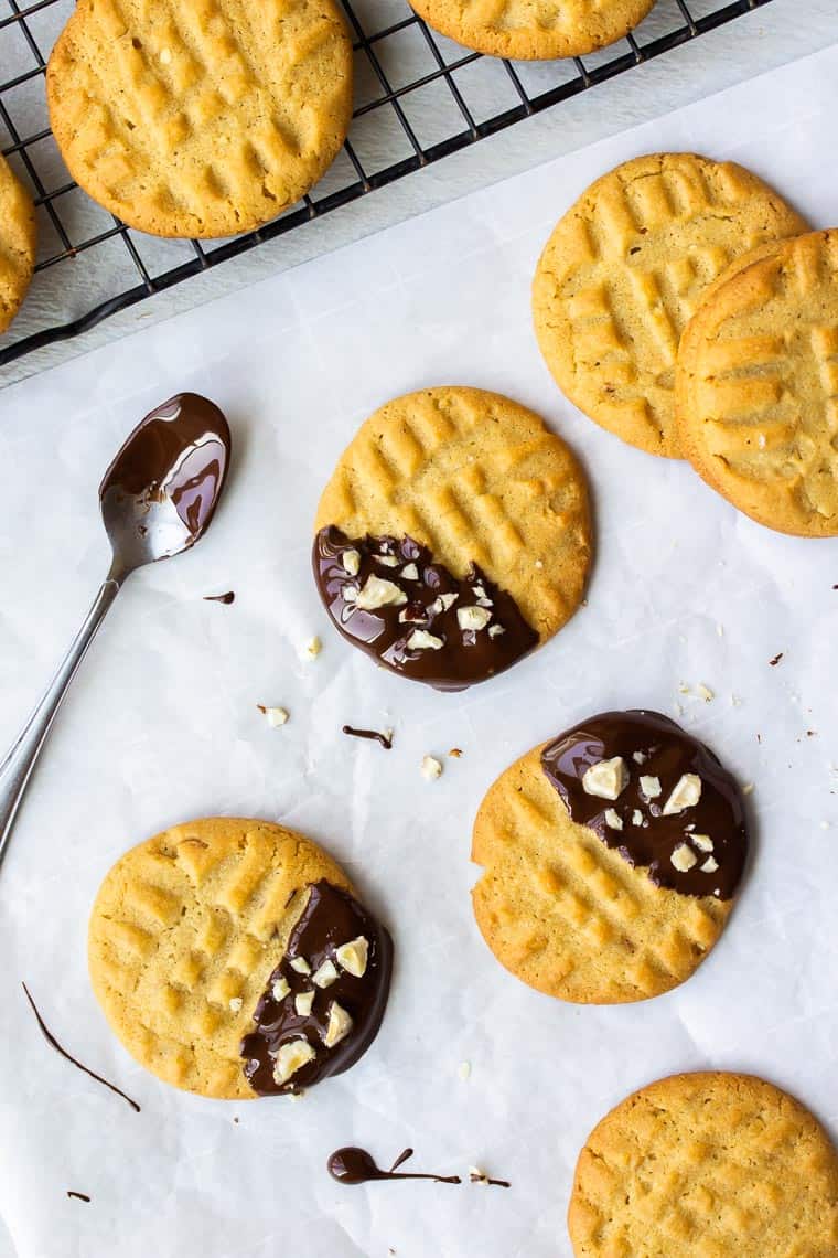 Overhead view of 3 chocolate dipped peanut butter cookies with a chocolate covered spoon, several undipped cookies. and a cooling rake with more peanut butter cookies on it over a white parchment paper with drizzled chocolate on it